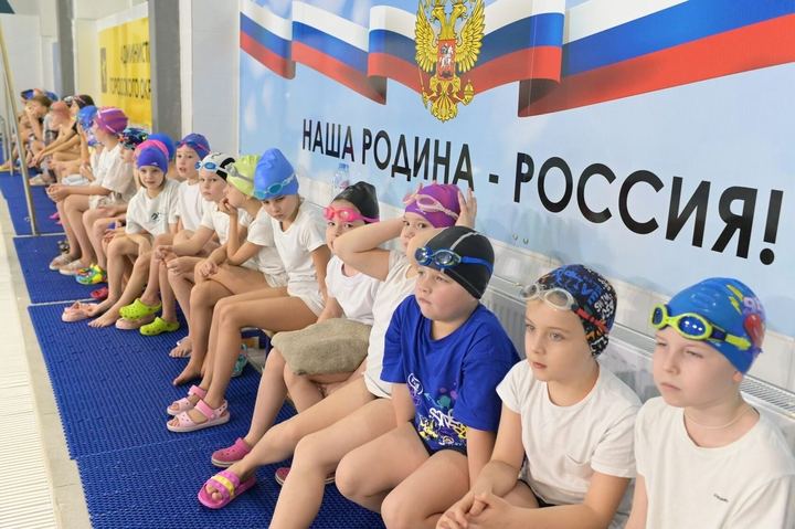 Юные пловцы на бортике бассейна перед тренировкой, на стене баннер с гербом и флагом России